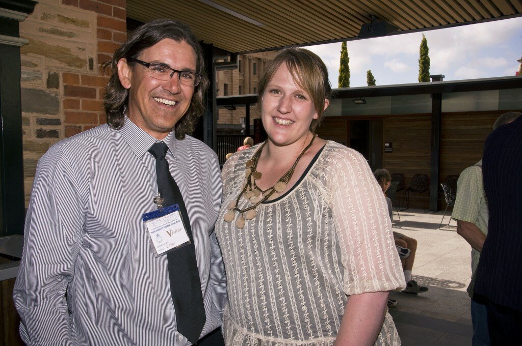 This is a photograph of Richard Bates and Bonnie Millen who are both looking in the direction of the camera and smiling. Mr Bates is standing on the left of the photograph. He is wearing a white and grey striped collared shirt with a black tie. He is wearing spectacles with a dark frame and has shoulder length curly brown hair. Ms Millen is on the right of the photograph. She is wearing a cream transparent blouse with a dark singlet underneath. She is also wearing a gold beaded necklaces made up of circular disk pendants. She has light brown hair with a fringe that is tied up.
