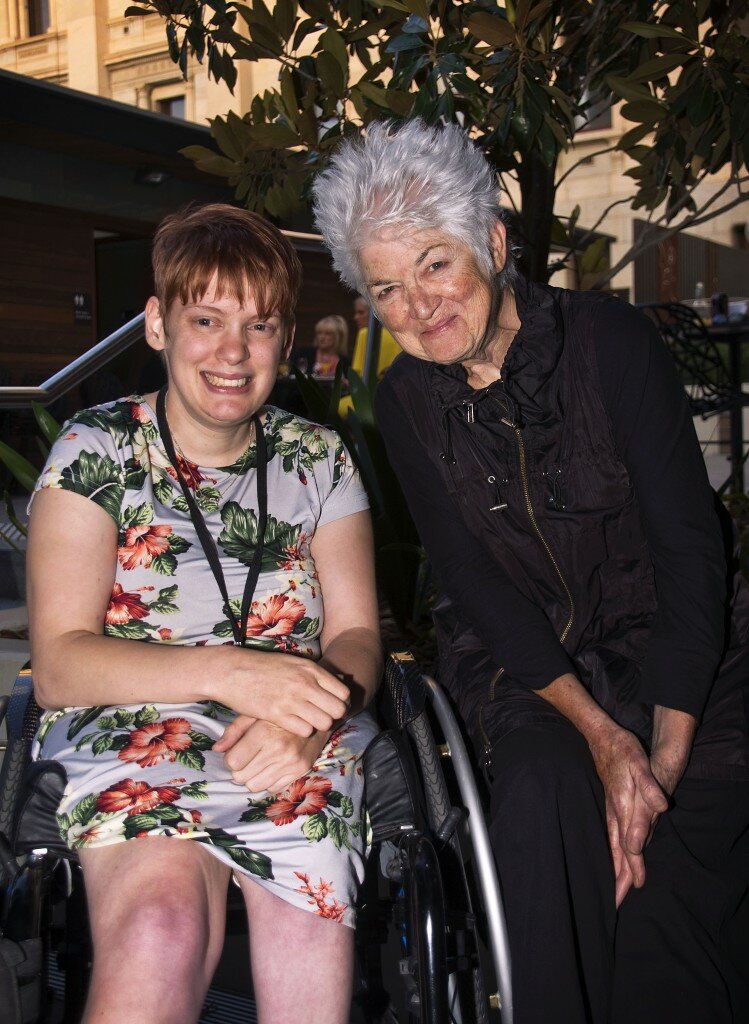 This is a photograph Kelly Vincent and PJ Rose. Ms Vincent is sitting in her manual wheelchair on the left hand side of the photograph. She is wearing a light blue dress with a pink, white, and green floral print featuring hibiscus and orchids. She is wearing a black land yard around her neck and has short red hair. She is smiling broadly and is looking in the direction of the camera. Ms Rose is sitting on the right hand side of the photograph. She is wearing a long sleeved dark purple top with a purple vest on top. She is also wearing very dark pants. She has short grey hair. Ms Rose is also smiling and looking in the direction of the camera. Directly behind Ms Vincent and Ms Rose is a small tree. There is also a building that is visible behind the tree made up of grey bricks and dark timber panels.