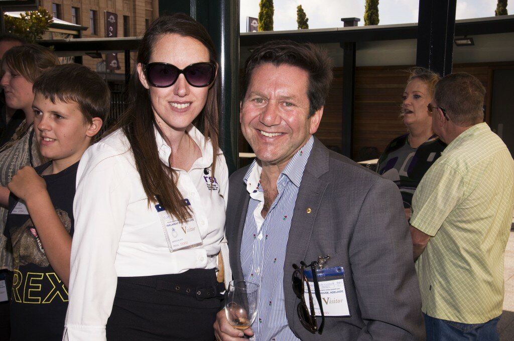 This is a photograph of Cathrin Gill and Freddie Brincat. Ms Gill is standing to the left of the photograph. She is wearing a Black skirt and a shite business shirt with an embroidered Royal Society for the Blind logo on the left in purple and black. She has long brown hair and is wearing large sunglasses with thick black rims and purple lenses. Mr Brincat is standing to the right of the photograph and is wearing a grey suit with a white business shirt with a thin blue stripe and tiny red rose design. He is holding wine glass containing a very small amount of white wine and a pair of sunglasses are hanging by one arm from his breast pocket. Ms Gill and Mr Brincat are hoth looking into the camera and smiling. in the background other guests at the end of year event can be seen standing and speaking with one another.