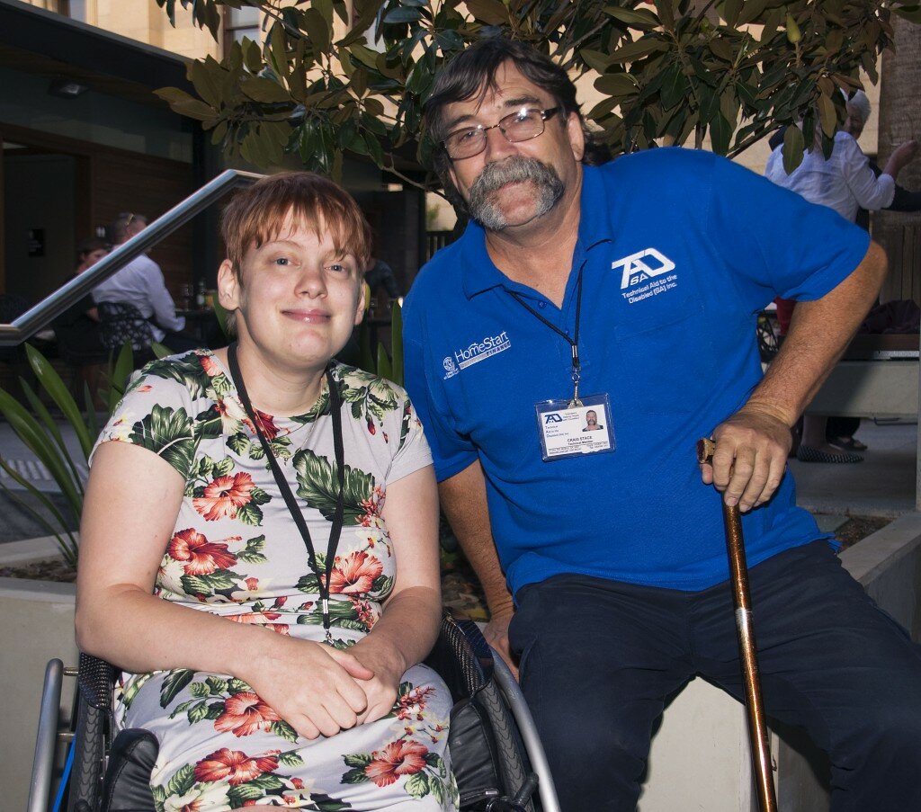 This is a photograph of Kelly VIncent and Craig Stace. Ms Vincent is sitting in her manual wheelchair on the left of the photograph. She is wearing a light blue dress with a pink, white, and green floral print featuring hibiscus and orchids. She is wearing a black land yard around her neck and has short red hair. She is smiling and is looking in the direction of the camera. Mr Stace is sitting on the right side of the photograph and is sitting on the edge of a cement garden bed. He is wearing a bright blue polo shirt with dark trousers. He is wearing spectacles with a metal frame and black land yard around his neck. He has dark brown hair and a long grey moustache. He is holding a cane in front of him. In the background is a small garden with a tree and small shrubs. Other guests can also be seen chatting behind them.