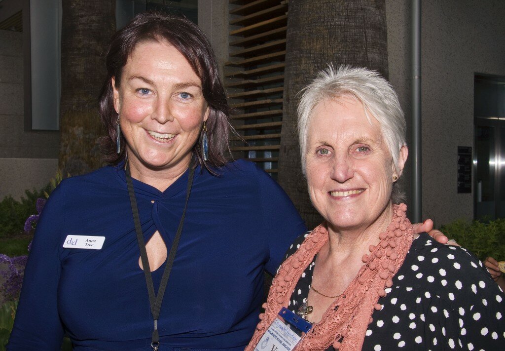 This is a photograph of Anna tree and Janne McMahon OAM, both of whom are looking directly into the camera and smiling. Only their heads and shoulders are visible in the photograph. Ms Tree is standing to the left of the photograph and is wearing a blue dress. Affixed to the dress is a name tag that reads "d4d Anna Tree" and around her neck hands an id card on a black lanyard. She has dark medium length hair and is wearing hanging earrings with long, blue ellipses. She has her left arm around Ms McMahon's shoulders. Ms McMahon is standing to the right of the photograph and is wearing a black dress with a large, white dot print, and a coral lace scarf with woollen baubles. She is wearing a thin gold necklace and gold stud earrings. She has short white hair and is wearing pink lipstick. In the background is a garden bed containing purple flowering plants and two very tall palm trees, whose think trunks extend up out of the upper margin of the picture.