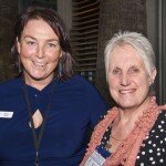 This is a photograph of Anna tree and Janne McMahon OAM, both of whom are looking directly into the camera and smiling. Only their heads and shoulders are visible in the photograph. Ms Tree is standing to the left of the photograph and is wearing a blue dress. Affixed to the dress is a name tag that reads "d4d Anna Tree" and around her neck hands an id card on a black lanyard. She has dark medium length hair and is wearing hanging earrings with long, blue ellipses. She has her left arm around Ms McMahon's shoulders. Ms McMahon is standing to the right of the photograph and is wearing a black dress with a large, white dot print, and a coral lace scarf with woollen baubles. She is wearing a thin gold necklace and gold stud earrings. She has short white hair and is wearing pink lipstick. In the background is a garden bed containing purple flowering plants and two very tall palm trees, whose think trunks extend up out of the upper margin of the picture.