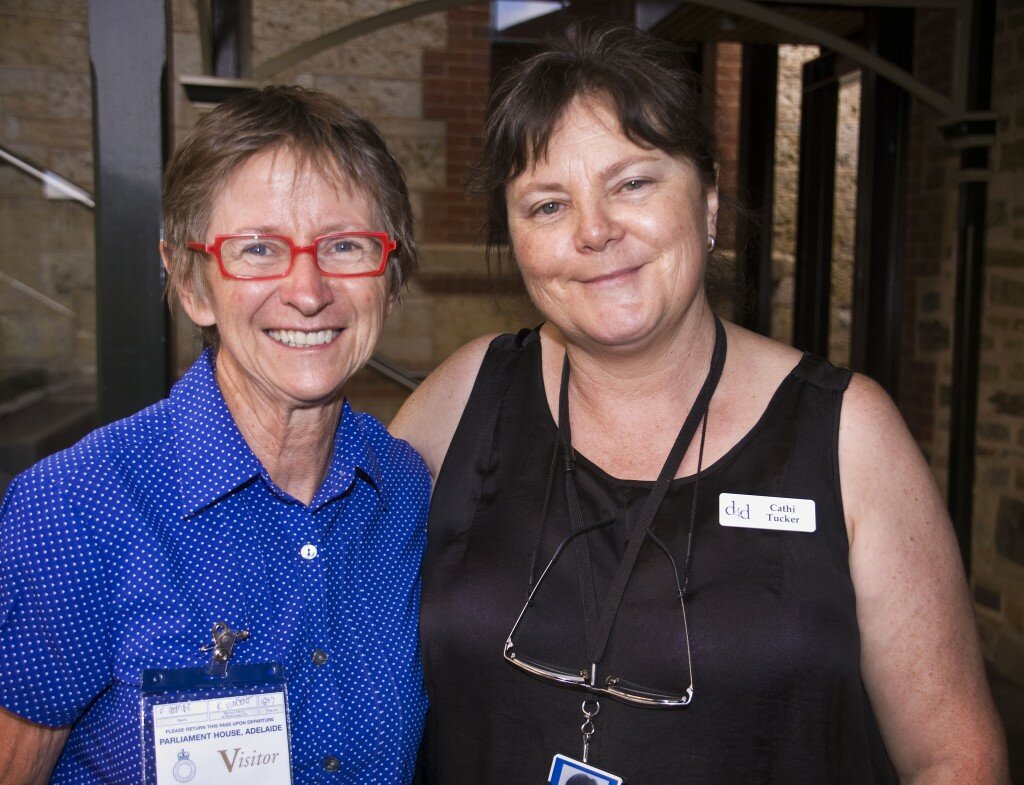 This is a photograph of Francis Coombe and Cathi Tucker. Ms Coombe is standing to the left of the photograph and looking directly in to the camera. Only her head and shoulders are visible. She is wearing a light blue button up shirt with short, capped sleeves with a fine, white, polka dot print. She has square spectacles with thick red frames. She has short brown hair and is smiling broadly. Ms Tucker is standing close to Ms Coombe, slightly to the right of the centre of the photograph. Only her head and shoulders are visible. She is wearing a black, sleeveless top and a nametag that reads "d4d Cathi Tucker". Around her neck hand an ID card on a black lanyard and a pair of spectacles with thin metal frames hung from a length of slim black cord. She is looking directly into the camera and is smiling. In the background of the photograph the blue stone wall of Old Parliament House is visible.