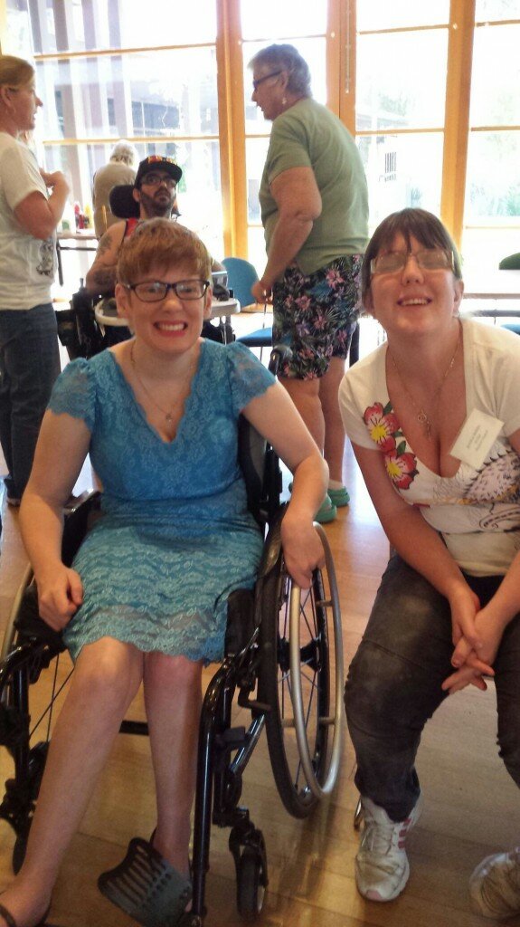 This is a photograph of Hon kelly Vincent MLC and Jess at the combined meeting of 5 local councils' access groups. Ms Vincent is in the left of the photograph and is seated in her manual wheelchair. She is wearing a light blue dress with a lace layer and has a thin gold chain with a small charm on it hanging around her neck. She has short red hair and is wearing pink lipstick and spectacles with thick black frames. Jess is seated in the right of the picture and is wearing blue denim jeans and a white t-shirt with a pink hibiscus print. She has dark hair pulled back into a ponytail and is wearing spectacles with thick brown frames. Both women are looking into the camera and smiling. In the background of the picture other attendees can be seen speaking with one another. 