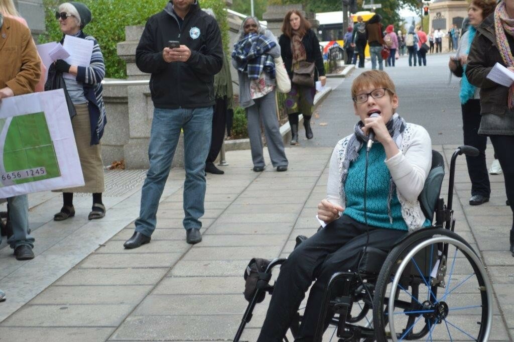 This is a photograph of Ms VIncent speaking at a rally opposing the use of compulsory income management of welfare payments. Ms VIncent is sitting in her manual wheelchair at the foot of the front steps of Parliament House. SHe is wearing black trousers, a teal shirt, a white cardigan, and a black and white scarf, and is speaking into a microphone she holds in her left hand. SHe has short red hair and is wearing spectacles with thick dark frames. Around her a group of people have gathered to listen to her speak.