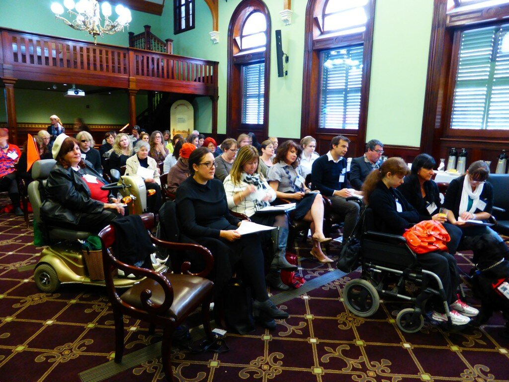 This is a photograph of the audience of the education forum. There are approximately 30 people in the audience who are all sitting down. The audience members are facing to the side of the photograph, facing in the direction of the presenters. The room has pale green walls with dark timber framed windows. Behind the audience in the top left of the photograph a is a dark timber balcony. There is also a golden chandelier in the top left of the photograph above the balcony. The carpet is a brown colour with a golden pattern.
