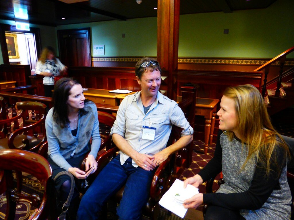 This is a photo of three audience members who attended the education forum. The audience members are sitting down and are facing each other. The photo was taken whilst the audience members were holding a conversation. In the background of the photograph the walls are a pale green and dark timber. There is also a pale wooden table behind the audience members. The carpet is brown with a gold print.