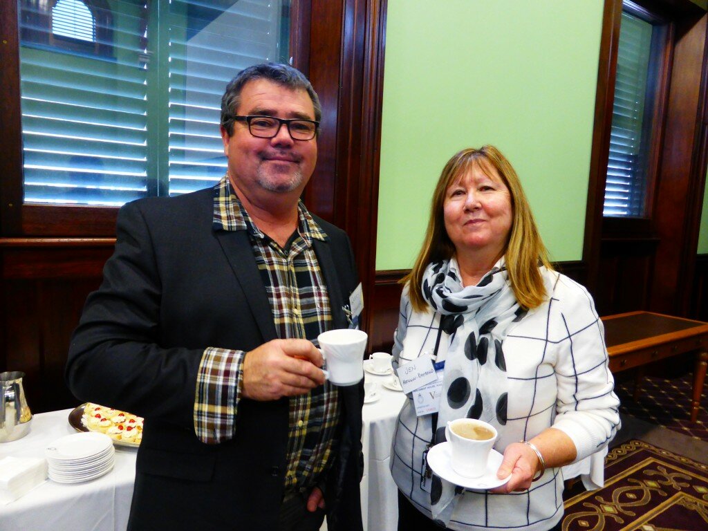 This a photograph of Garry Connor and Jen Mathwin-Raymond. Mr Connor is on the left side of the photograph and is facing the camera. He is smiling and holding a cup of tea. He is wearing a dark tartan shirt with a black blazer over the top. He has short black hair. Ms Matthwin-Raymond is on the right of the photograph and is facing the camera. She is also smiling and holding a cup of tea. She is wearing a white long sleaved top with black lines. She is also wearing a white scarf with black polka dots. She is shoulder length blonde hair. In the background the walls are pale green. There are two large windows which have dark timber frames. In the bottom left of the photograph a table can be seen with plates and scones displayed. The carpet is brown with a gold pattern