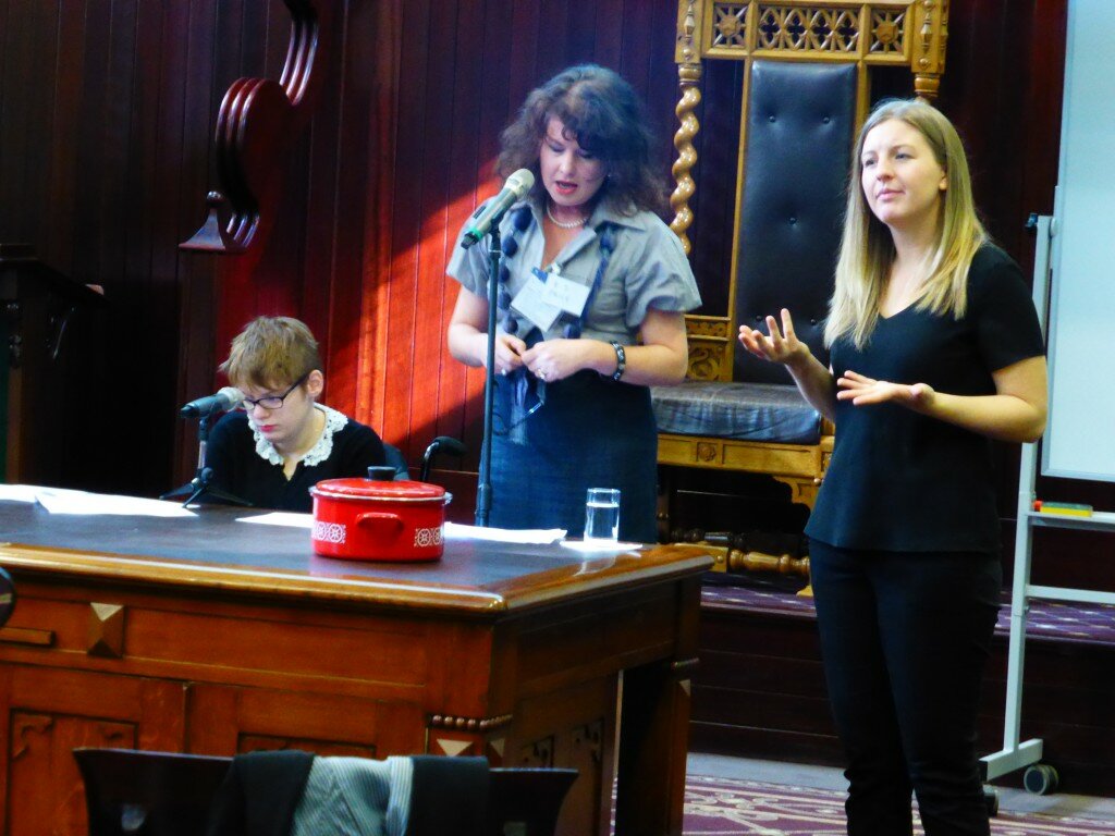This is a photograph of Kelly Vincent, B-J Price and a sign language interpreter addressing the education forum. Ms Vincent is seated behind a desk to the left of the photograph in her manual wheelchair. She is looking away from the camera and is looking down whilst she listens to Ms Price speak. She is wearing a black cardigan with a white floral lace collar. She is wearing spectacles with a dark frame and bright red lipstick. She has short, red hair. Ms Price is standing in the middle of the photograph and is also standing behind a desk. She is looking down and is speaking into a microphone. Ms Price is wearing a light purple blouse and a dark navy skirt. She is also wearing dark purple scarf that is made up of pom-poms. She is holding her spectacles in her hands. She has brown shoulder length hair with a fringe. The sign language interpreter is standing to the right of the photograph. She is signing while Ms Price speaks. She is wearing a black t-shirt and pants and has long, blonde hair. In the background of the photograph is a dark timber stage. The stage has a large chair on it with a light wooden frame and leather cushioning. The carpet can also be seen on the stage with is a burgundy colour with a gold square pattern.