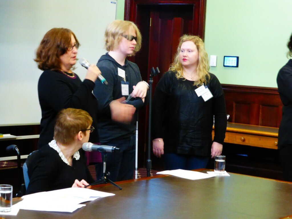 This is a photograph of Kelly Vincent, Mimi Everett, Charlie Everett and Eloise Everett. Ms Vincent is sitting behind a desk to the left of the photograph. She is facing away from the camera and is facing Ms Everett while she speaks. She is wearing a black cardigan with a white floral lace collar. She is wearing spectacles with a dark frame and has short, red hair. Ms Everett is standing to the left of the photograph behind Ms Vincent. She is facing away from the camera in the direction of the forum audience and is speaking into a microphone. She is wearing a black long sleaved top with a purple stripe at the bottom. She is also wearing a gold and purple necklace and spectacles with a dark frame. She has shoulder length brown hair. Standing beside Ms Everett in the centre of the photograph are her children Charlie Everett and Eloise Everett.Charlie is wearing a grey hooded jumper and dark glasses and is holding a cane. Eloise is wearing a long sleeved black top and denim jeans. She has shoulder length blonde hair and is wearing red lipstick. In the background behind Ms Everett is a blank white screen. To the right of the photograph is a timber door which is surrounded by a pale green wall.