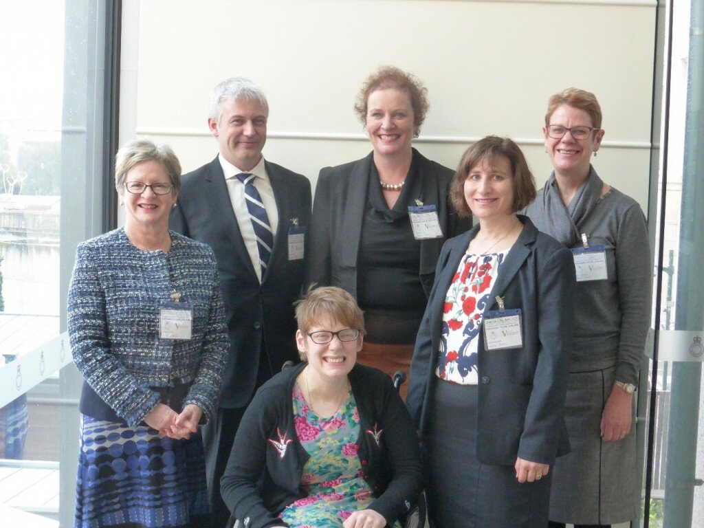 This is a photograph of Kelly Vincent with a group of five Speech Pathology presenters. Ms Vincent is at the front of the group, sitting in her manual wheelchair. There are five presenters standing behind Ms Vincent. Everyone in the photograph is facing the camera and smiling.