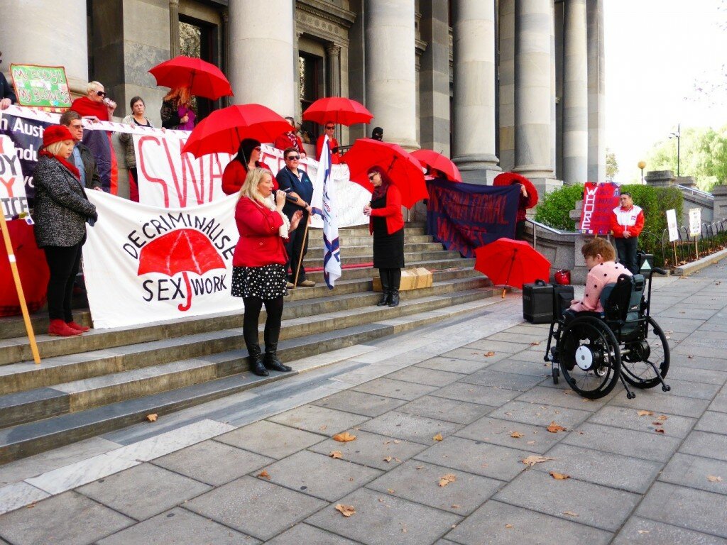This is a photograph of the International Whore's Day Rally, that took place in South Australia on the steps of parliament. Kelly Vincent is on the right hand side of the photograph, she is facing the rally attendees who are standing on the steps of parliament. Tammy Franks is also facing Ms Vincent, she is speaking into a microphone to make a speech at the rally. Most of the attendees are dressed in bright red and holding banners or umbrellas.