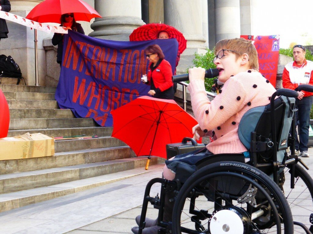 This is a photograph of Kelly Vincent speaking at the International Whore's Day Rally. She is sitting to the right of the photograph and is addressing the rally attendees. She is speaking into a microphone and is sitting in her manual wheelchair.