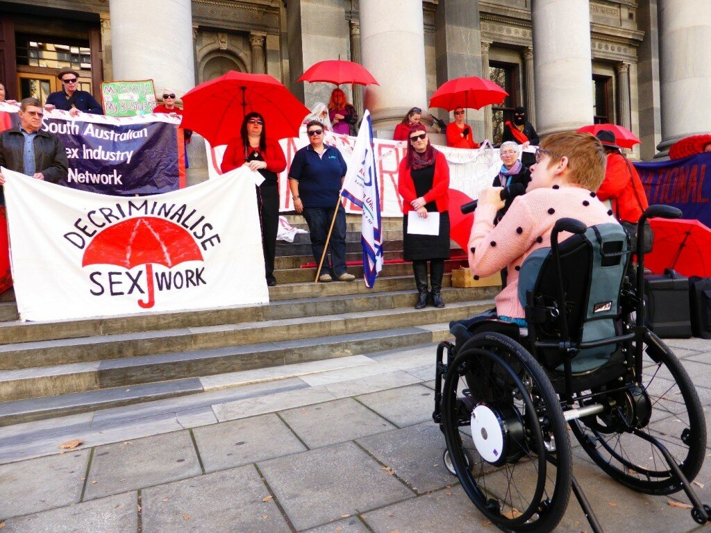 This is a photo of Kelly Vincent addressing the International Whore's Day Rally that was held in South Australia on the steps of parliament house. Ms Vincent is facing with her back to the camera. She is speaking into a microphone and is sitting in her manual wheelchair. There are 11 rally attendees that are visible in the photograph who are standing on the steps of parliament. Many attendees are wearing red and holding banners and umbrellas.