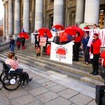 This is a photograph of Kelly Vincent speaking at the International Whore's Day Rally. The photograph has been taken on the steps of Parliament House in South Australia. Kelly Vincent is facing away from the camera and is speaking in to a microphone. Ms Vincent is speaking to a crowd of around 20 people, many of which are wearing red or holding bright red umbrellas. Many are also holding decriminalise sex work banners.