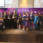 This is a photograph of Kelly Vincent and 11 others at the Workability Asia Conference. The group are standing in a line on stage with Ms Vincent sitting in her manual wheelchair on the left side of the photograph. Everyone on the stage is smiling and is looking to the left of the photograph towards the audience which is out of frame. Everyone is dressed in a smart business manner. In the background of the stage is a purple backdrop. There is also a sign to the left of Ms Vincent that says "welcome".