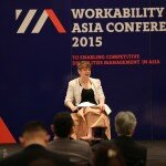 This is a photograph of Kelly Vincent speaking at the Workability Asia Conference. Ms Vincent is in the centre of the photograph, sitting in her manual wheelchair. Ms Vincent is looking to the right of the camera in the direction of the audience. Her mouth is slightly open as she is making a speech. Ms Vincent is wearing a black dress with a white lace blazer and has a beige blanket draped on her lap. She is holding on to her notes which are sitting on her lap and she has short red hair. Ms Vincent is on a stage and a few audience members can be seen infront of the camera. Behind Ms Vincent is a big dark blue sign that says 'Workability Asia Conference 2015' in white font.