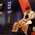 This is a photograph of Kelly Vincent speaking at the Workability Asia Conference. Ms Vincent is sitting to the right of the photograph in her manual wheelchair. Ms Vincent is looking down in the direction of her speech which is sitting in her lap. Ms Vincent is wearing a black dress with a white lace blazer and has a beige blanket draped over her lap. Ms Vincent also had a small microphone taped to her right cheek and has short red hair. In the background of the photograph is a big sign that says ' Together we can make a difference'. The sign is black and dark blue striped with white and red text.