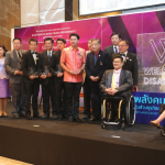 This is a photograph of Kelly Vincent and 12 other speakers at the Workability Asia Conference. The group are up on a stage and are looking to the left of the camera in the direction of the audience which is not in frame. Ms Vincent is sitting in her manual wheelchair on the right of the photograph. On the left of Ms Vincent is a sign that says ' Welcome'. The 12 speakers are on the other side of the sign, situated in the left and centre of the photograph. Everyone in the photograph is dressed in a smart business manner and are smiling.