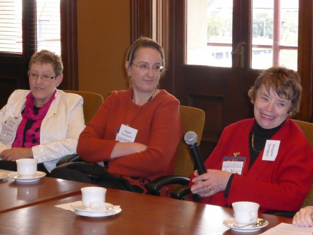 This is a photograph of Christine Russell, Bogda Koczwara and Margaret Springgay. Ms Russell is sitting to the left of the photograph. She is wearing a white blazer with a black blouse and bright pink scarf. She is wearing small spectacles with a metal frame and has short blonde hair. Ms Koczwara is sitting in the centre of the photograph. She is wearing an orange cardigan with a black floral print. She is also wearing spectacles and has long brown hair that is plaited. Ms Springgay is sitting to the right of the photograph. She is wearing a black top with a bright red cardigan on top. She has short brown hair and is holding a microphone. The women in the photograph are looking to the right of the camera in the direction of someone speaking who is out of frame. They are all siting at a table and have three cups of tea in front of them.