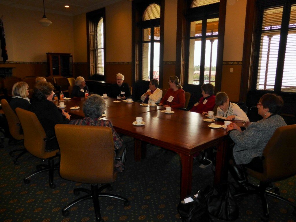 This is a photograph of all of the morning tea attendees sitting around a table. The group are looking in the direction of Rosemary Crowley who was speaking when the photograph was taken. In the background of the photograph are very large windows and a door that goes out to a balcony. The group are all siting at a table with cups of tea and plates in front of them.
