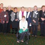 This is a photograph of all of the women that attended the morning tea. From left to right - Valmai Hankel, Bogda Koczwara, Monica Oliphant, Rosemary Crowley, Jane Lemon, Christine Russell, Yvonne Hill, Lesley Hawkins, Tracey Murphy, Margaret Springgay. Kelly Vincent is sitting in her manual wheelchair in front of the group. They are all smiling and looking in the direction of the camera.