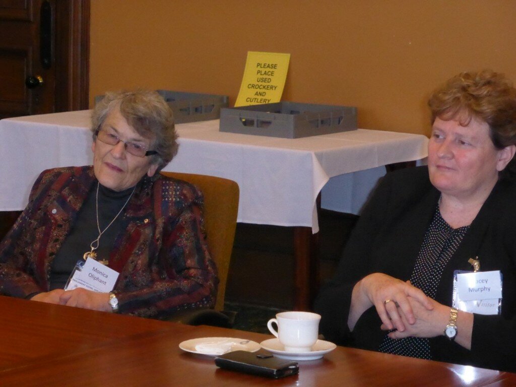 This is a photograph of Monica Oliphant and Tracey Murphy. Ms Oliphant is sitting to the left of the photograph. She is wearing a grey top with a colourful striped jacket made up of orange, blue, red, green, yellow, purple and grey. She is wearing small spectacles and has short grey hair. Ms Murphy is sitting to the right of the photograph. She is wearing a black and white polka dot patterned blouse with a black blazer. She has short blonde hair. Both Ms Oliphant and Ms Murphy are sitting at a table with a cup of tea and a plate in front of them.