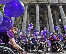 This is a photo of Kelly Vincent and the crowd at the Disability Pride Parade 2016. In it, the crowd are mostly wearing purple and are holding purple ballons. The photo was taken out the front of Parliament House, and Parliament House can be seen in the background of the photo