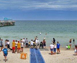 This is a photo of the Accessible Beach Day at Henley Beach. In the photo, the accessible beach mat appears in the centre of the photo, and is blue against the faint yellow sand. There are lots of people pictured on the beach mat. There are lifeguards in the photo, and the water of the sea fills the background of the image.